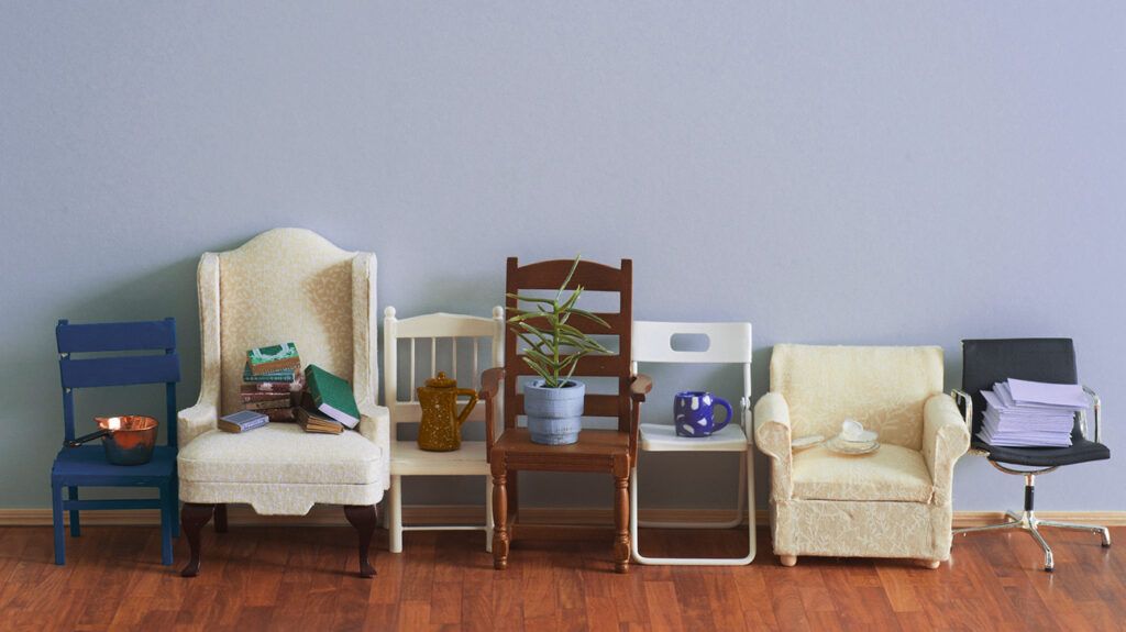 row of seven chairs against a wall with stacks of books papers plants and pots 