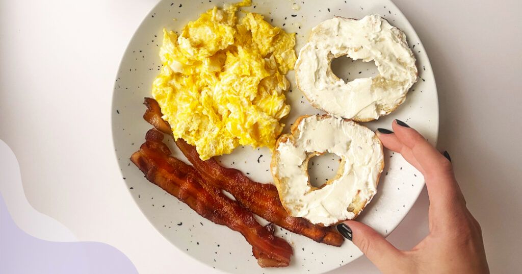 A plate with scrambled eggs, a bagel with cream cheese and two strips of bacon.