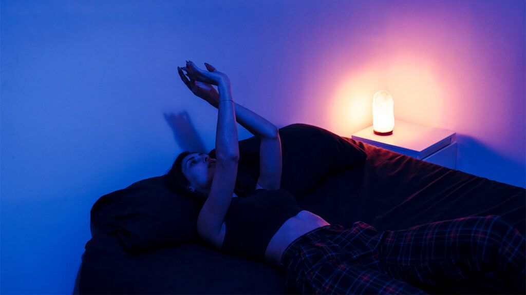 female laying in bed with their hands in the air making shadow shapes and a dim nightlight on a bedside table. They could be contemplating whether sleep anxiety medications to help 