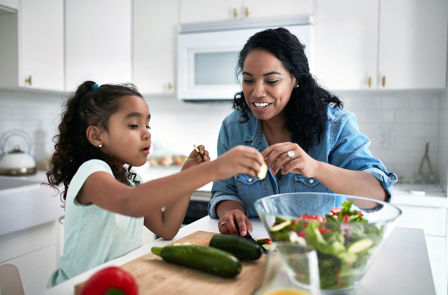 Woman and young girl smiling making salad together in bright while kitchen