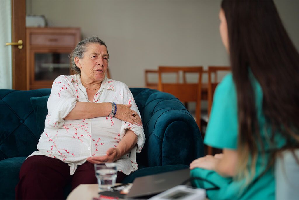 Older adult female sitting in an armchair holding her left arm discussing shingles vaccine side effects with a healthcare professional sitting opposite