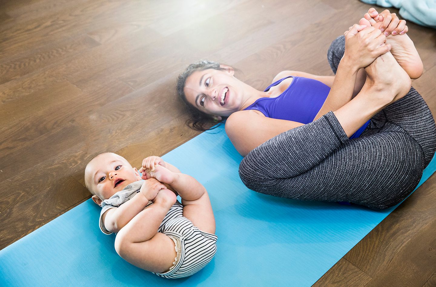 Woman doing stretches on her back on yoga mat looking at smiling at baby next to her in same pose holding legs