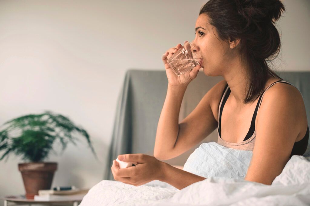 Female sitting up in bed, drinking from a glass of water while taking Zoloft.