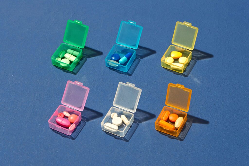 Six different pill pots in different colors. Each pot has pills that are the same color as the pot. The pots are transparent and in two rows of three. Top row is green, blue, and yellow. Second row is pink, white, and orange. These could be antipsychotic drugs for schizophrenia in a particular order.