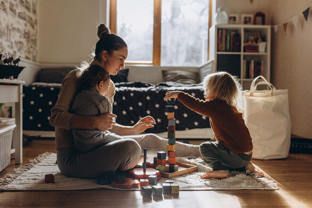 Adult female sitting crosslegged on the floor with a young child on her lap and another in front of her. Between them they are stacking some multi colored bricks to possibly calm genetic anxiety 