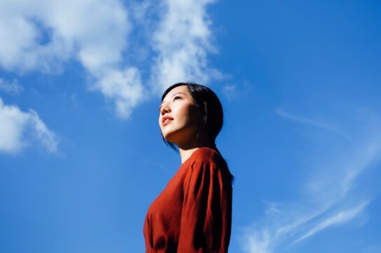 Adult female standing in looking into the distance with a background of blue sky with a few wispy clouds. The bright sunshiny day helps to get vitamin d for depression