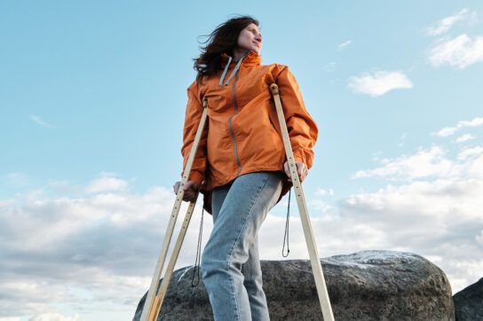 Adult female on a hike in nature, standing in front of a large flat oval rock using crutches, depicting types of MS and how they can still allow fun, physical activity