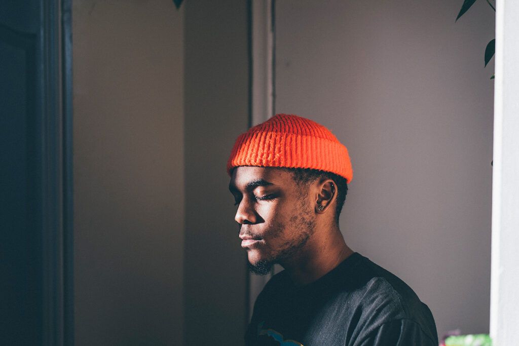 Young Black man wearing orange knit cap looking toward window with eyes closed in thought