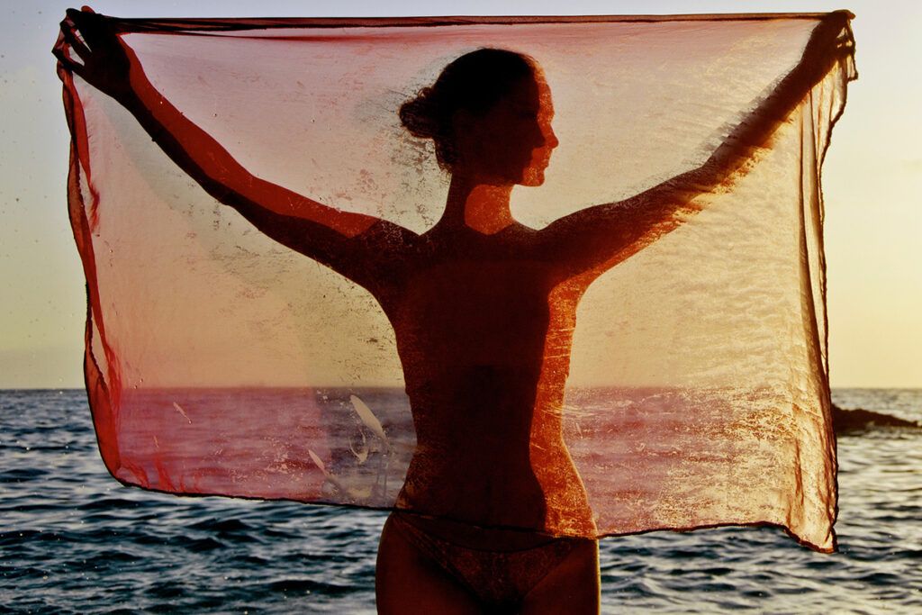 A person standing in front of the sea holding up orange translucent fabric.