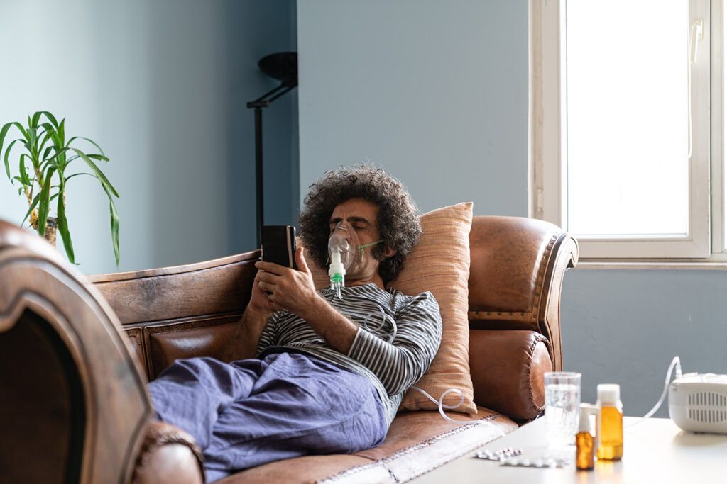 A person lying on a couch looking at their phone and using a nebulizer, which is a treatment method for COPD.