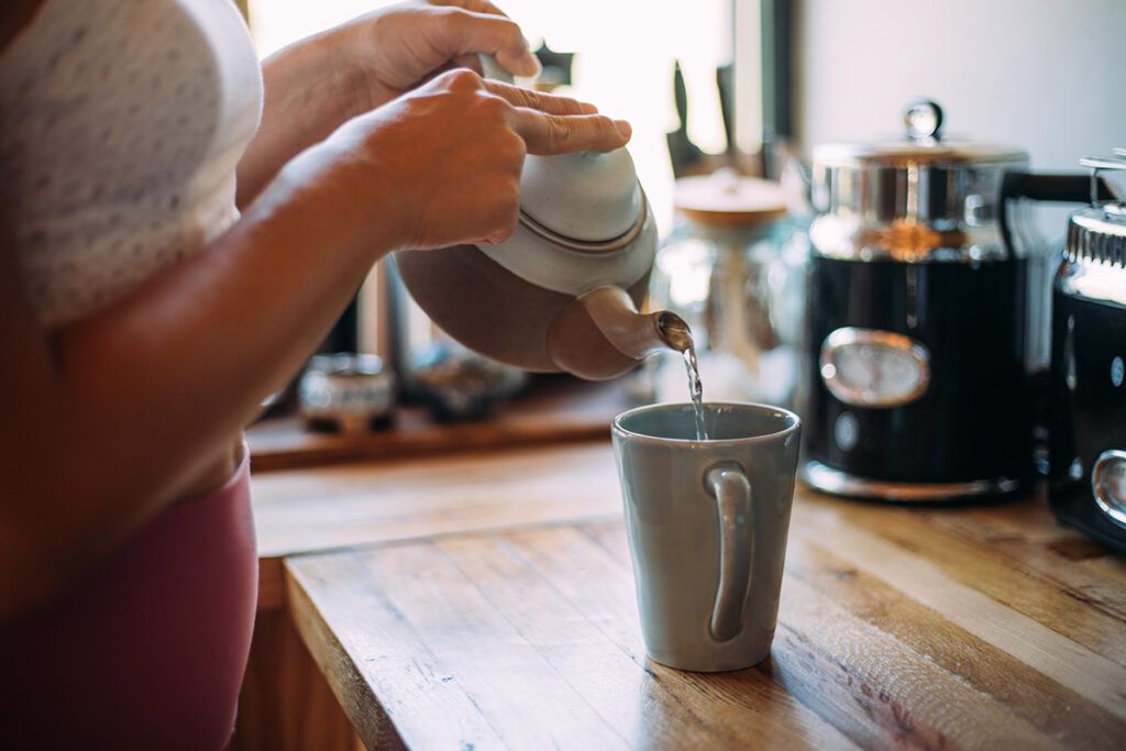 Close up of a person poring tea into a tall mug on a wooden kitchen worktop depicting natural remedies for osteoarthritis