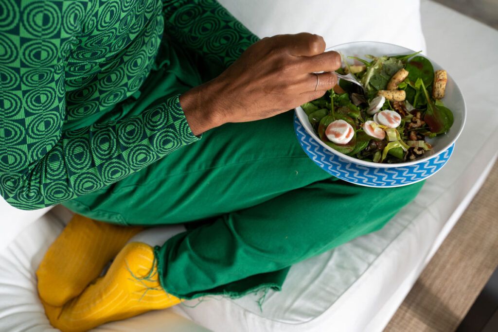 A person shown from the neck down sitting on a couch eating a salad, an example of a low sodium diet for hypertension.