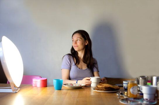 Female sitting at a table with several dishes surrounding her, while facing a light therapy lamp.