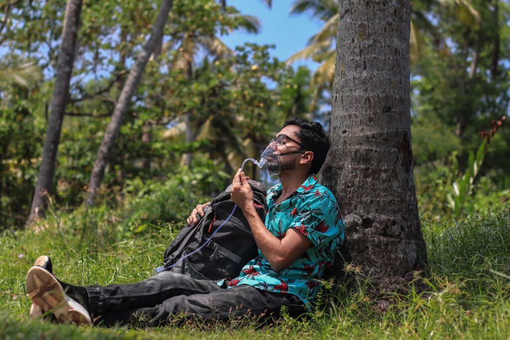 An adult sitting outside against a tree with an oxygen mask on his face, breathing in extra oxygen.