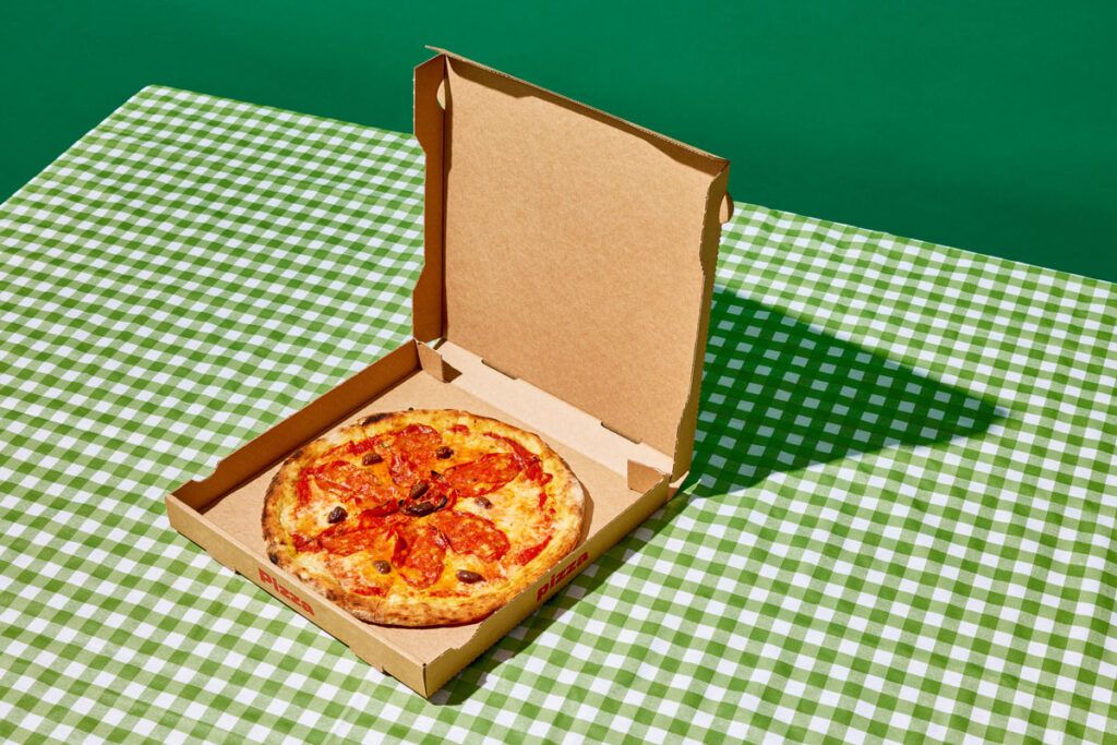 A whole pizza in a box, sitting on a green and white checked table cloth.