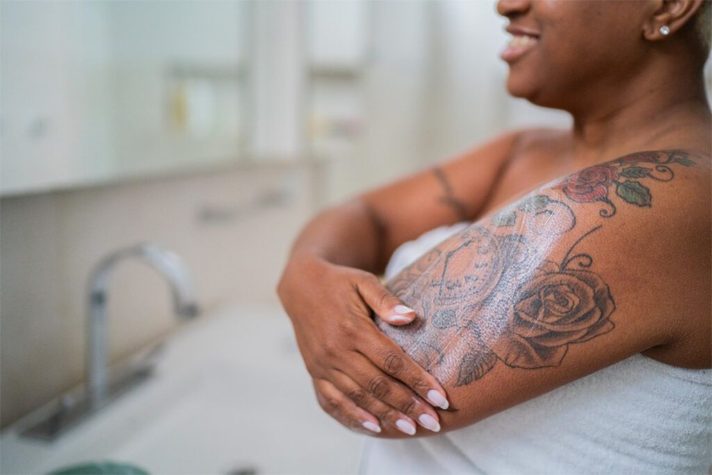 A Black person with a tattoo applying cream to their arm, representing eczema on Black skin and how it may look.