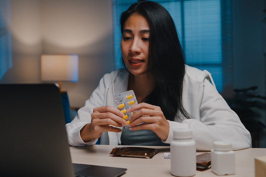 A young person sitting in front of a laptop and some medication bottles holding a packet of nausea medication.