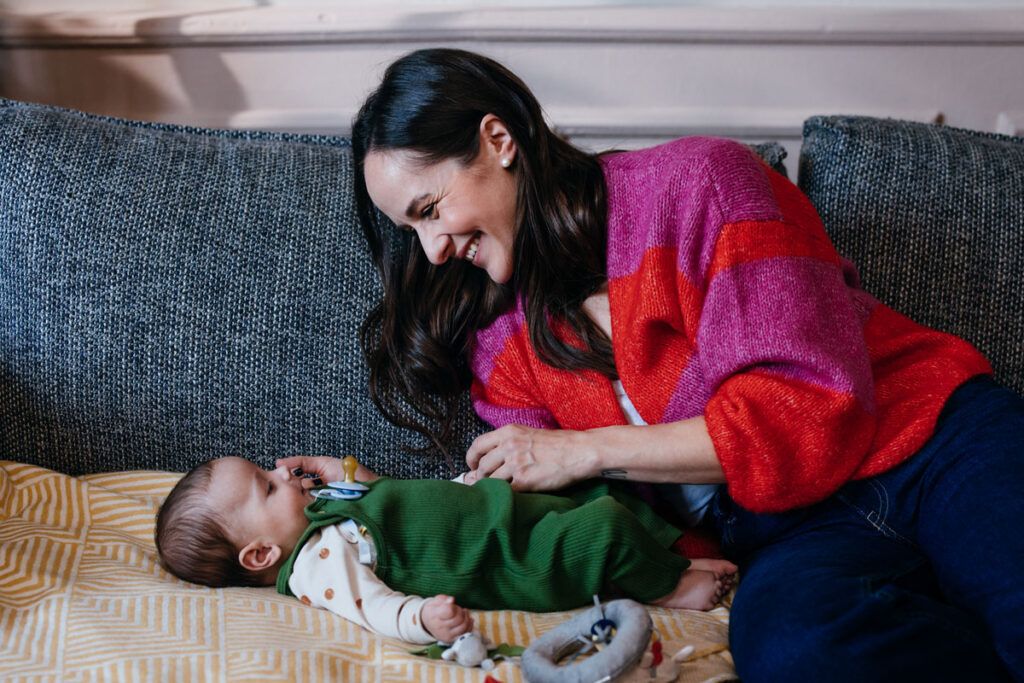 Female smiling and leaning over a baby who is laying down on a couch.