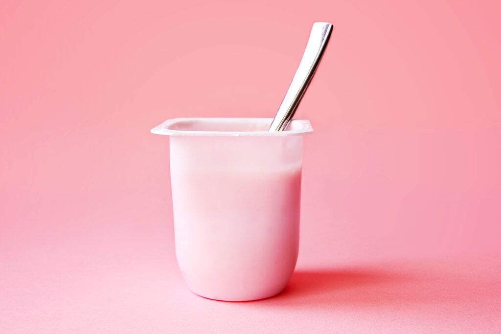 A yogurt that contains probiotics for eczema with a spoon in it against a pink background.