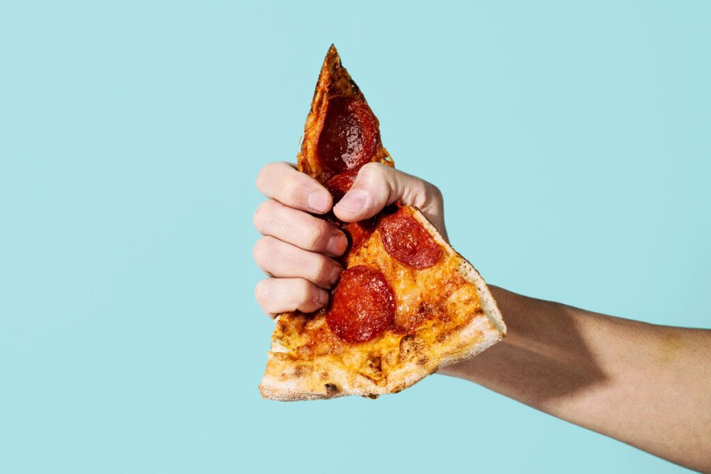 A hand clenching a slice of oily pepperoni pizza, which is a cause of acid reflux and shortness of breath.