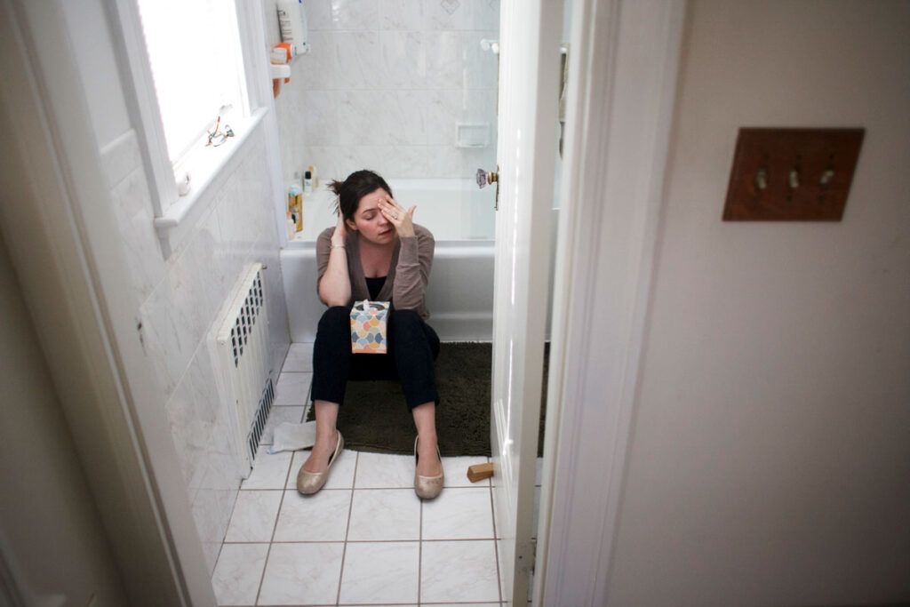 A woman experiencing nausea from acid reflux sitting on a bathroom floor holding tissues.