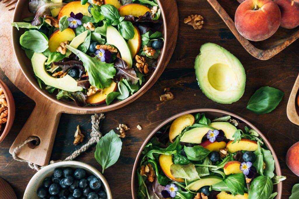 Two bowls of salad consisting of kale, avocados, peaches, walnuts, and blueberries.