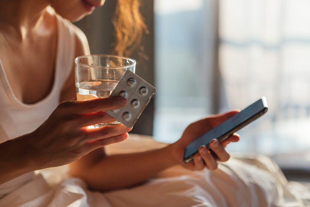 Close up of an adult female sitting in bed with morning sunlight coming through the window, holding a blister pack of pills and a small glass of water in one hand and looking down at her phone that's in the other hand checking when to take atorvastatin morning or night