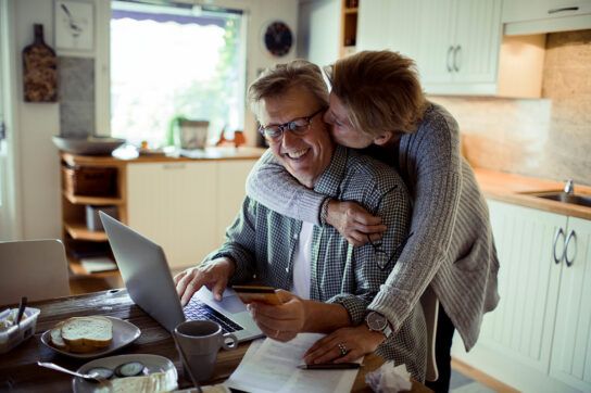 Older adult couple in a kitchen with a male sitting at table looking at a laptop and smiling as the female hug him around his neck from behind and kisses his cheek as they look for ED cures for seniors