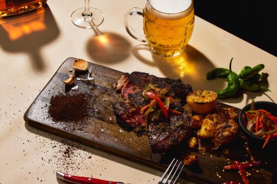 A steak on a serving plank with chillis and beer next to it. These are purine-rich foods that affect gout.