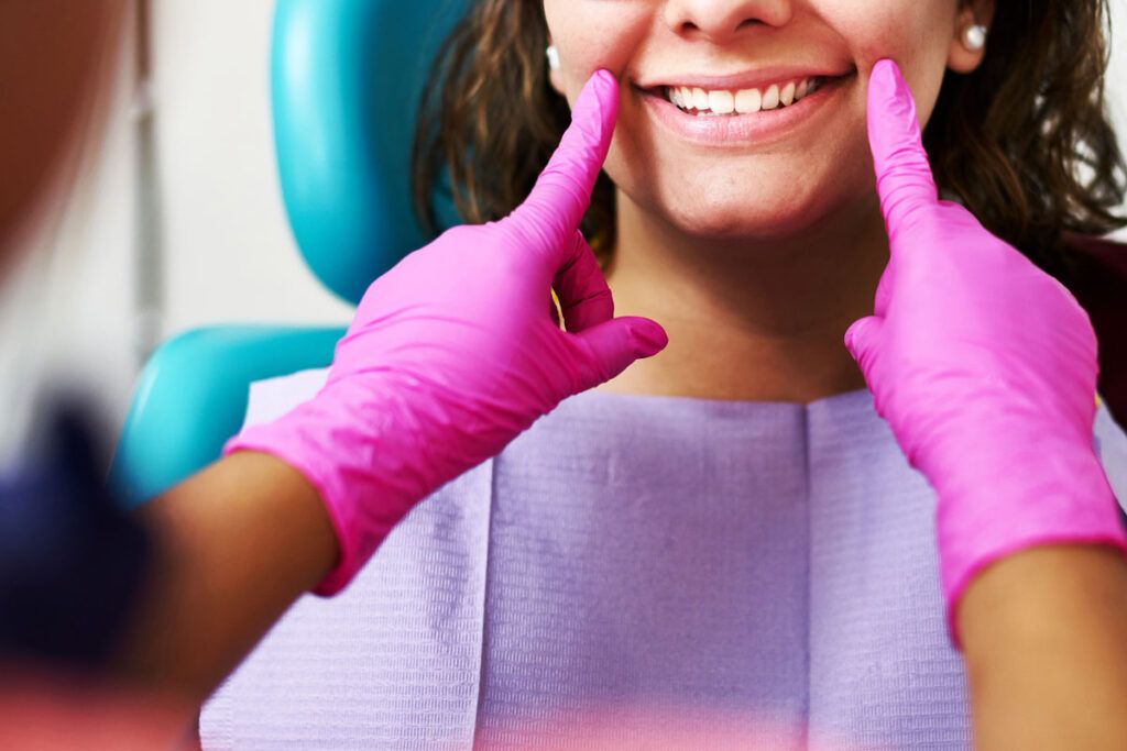 Person sitting in a dental chair as a healthcare professional wearing pink gloves touches their mouth. 