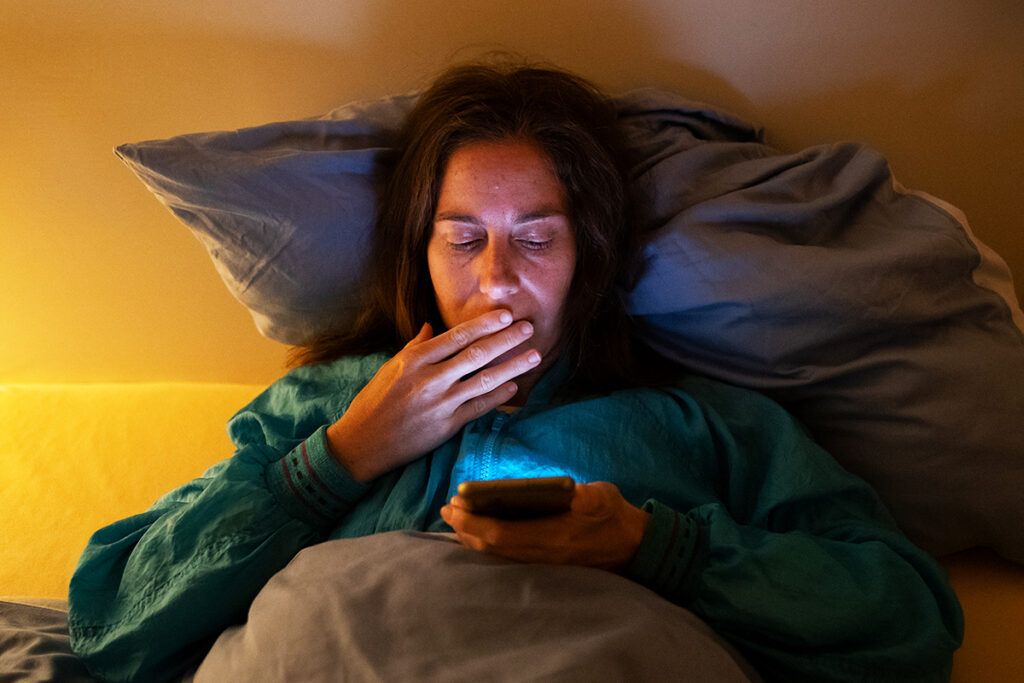 Adult laying in bed with a night light on and yawning looking at a mobile device wondering how to manage anxiety at night
