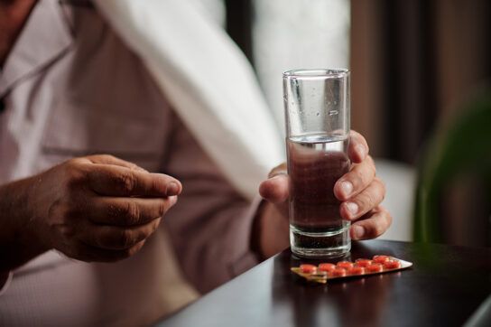 Close up of a person holding a tall glass of water in one hand and a pill in the other with a blister pill pack in front of them on a table depicting Lexapro side effects first week