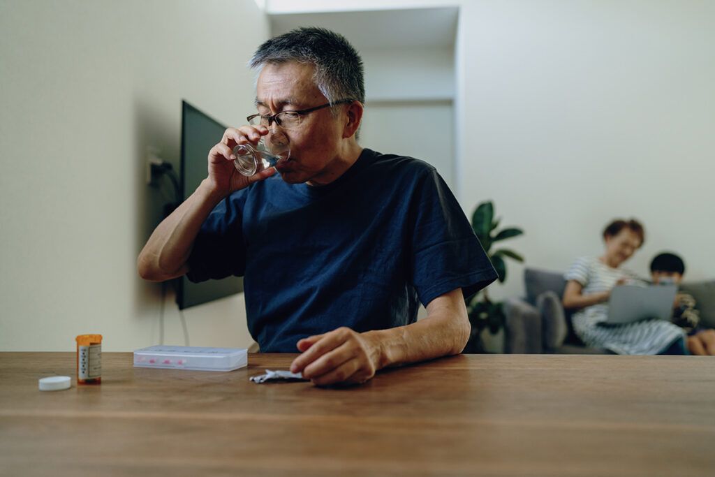 Man at kitchen table taking medications from pill box, possibly statins