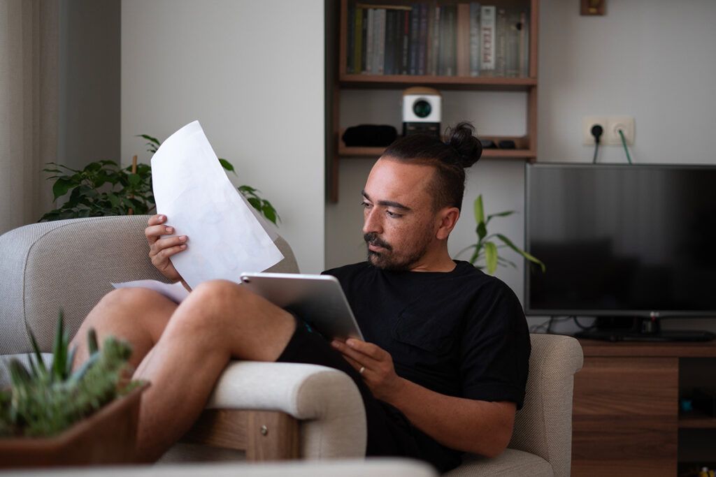 Adult male sitting comfortably at home looking through paperwork and a tablet device after thinking should I get an high deductible health plan