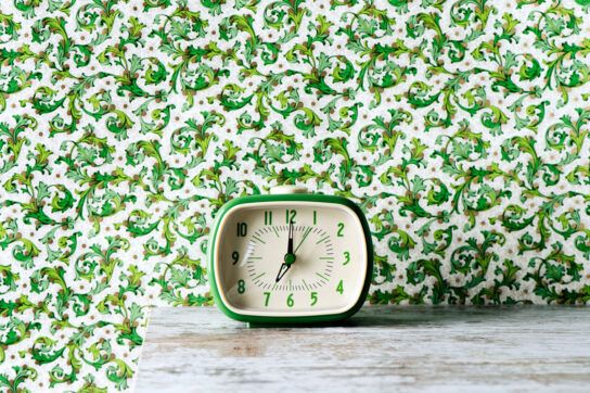 Wallpaper with a bust green vine pattern and a small green alarm clock on a stripped down wooden table that you could set once you have figured the best time to take Lexapro for anxiety depending on how it affects your body