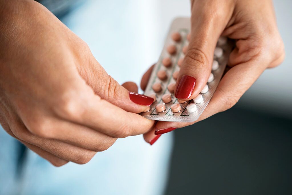 A person wearing red nail polish holding a packet of birth control pills, wondering about the long-term side effects.