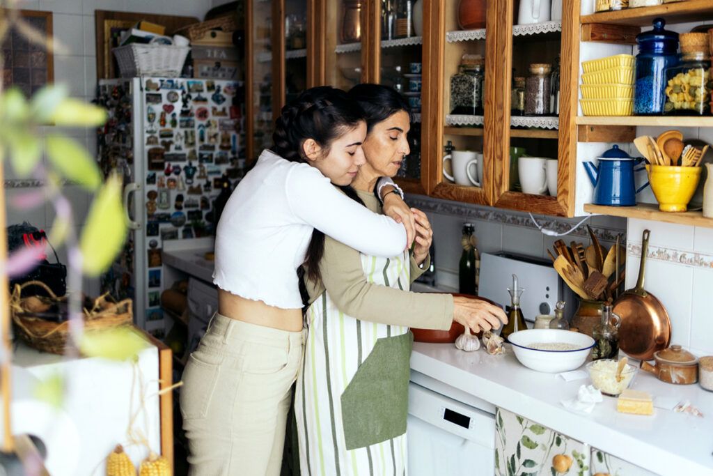 An older adult cooking in a kitchen, while her daughter hugs her from behind. She may be taking memory loss medications.