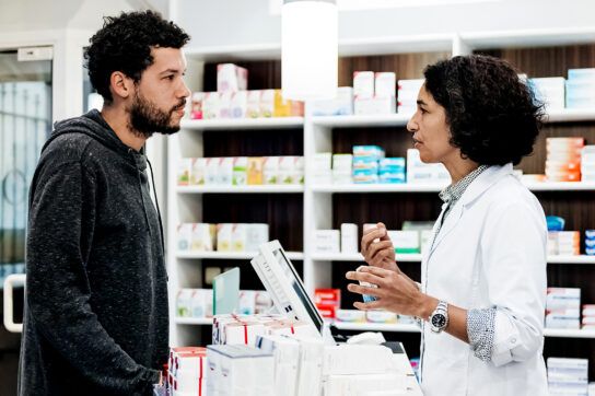 Person standing at a pharmacy counter speaking with a pharmacist to depict questions about using Optum Perks discount card with health insurance.