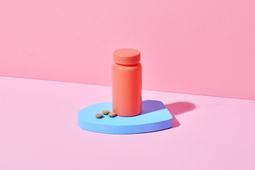A pink pill bottle on a blue board against a pink background. There are some anticholinergics in front of it.