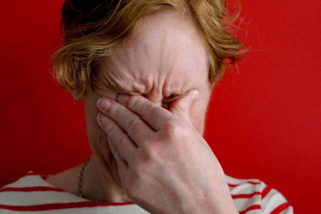A person shown from the neck up against a red background rubbing their eyes because they have gritty eyes.