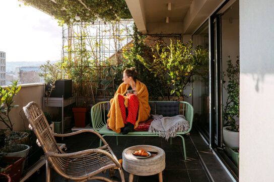 A young adult wrapped in a blanket with a cup of tea, sitting on a bench on a cosy-looking balcony. This represents how Xanax feels and its calming effect.