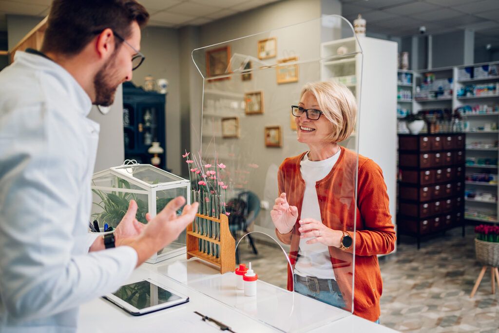 Female standing behind a glass screen and asking a pharmacist questions.