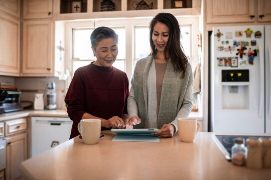 Two adult females standing at a breakfast bar in a home kitchen looking at a tablet device together searching for how do you qualify for Medicaid