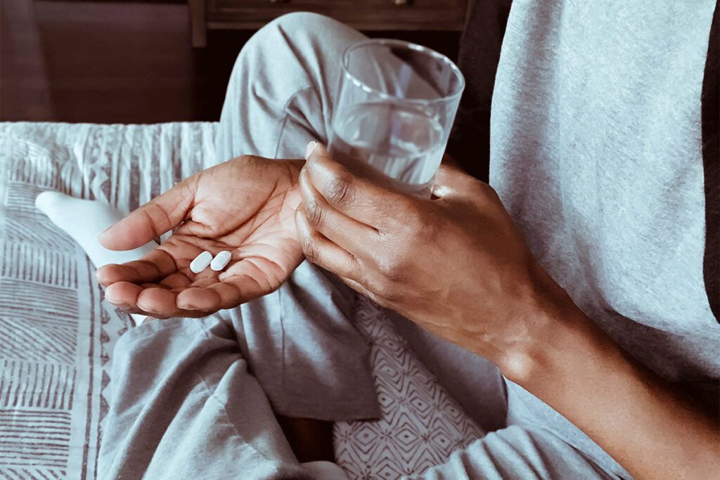 Person sitting with their legs crossed, holding a glass of water and 2 pills, to depict the question: should I take antibiotics?