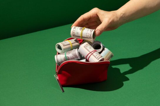 Small zipper pouch with rolls of money spilling out and a hand placing another roll of money on top depicting why are prescription drugs so expensive