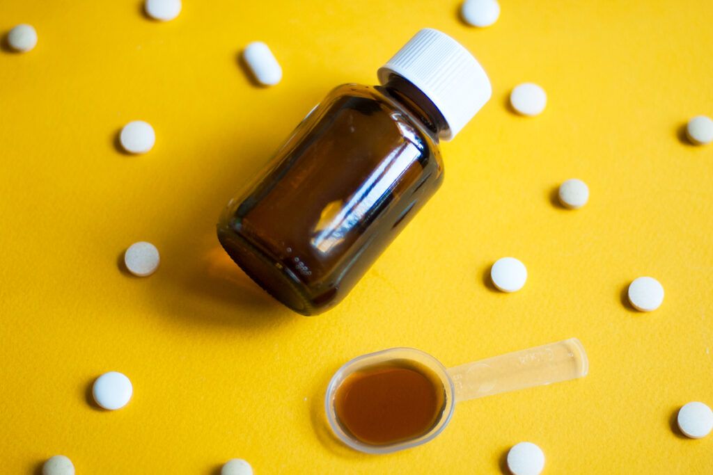 Yellow surface with several pills scattered across it and a spoon of cough syrup, to depict over-the-counter medication for flu.