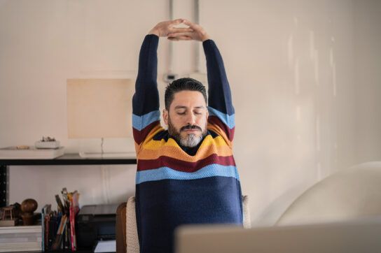 Adult male sitting at a home office desk and stretching his arms above his head with his hands joined to try and avoid stiff joints