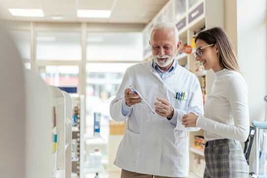 Older male adult pharmacist helping a female in a pharmacy showing her how to compare drug prices