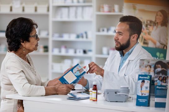 Adult male healthcare professionals speaking to an older female adult in a pharmacy setting discussing prescription card vs. insurance card