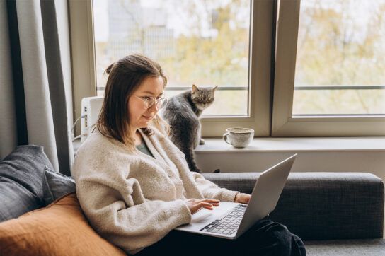 Adult female sitting in a living room chair next to a window with a cat sitting next to her on the window ledge and a cup of coffee next to the cat. The female is looking on her laptop possibly asking can you use HSA to pay for prescriptions